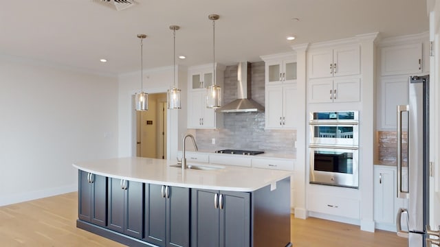 kitchen with wall chimney range hood, sink, appliances with stainless steel finishes, white cabinets, and a center island with sink