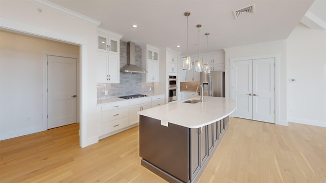 kitchen featuring sink, stainless steel appliances, an island with sink, decorative light fixtures, and wall chimney exhaust hood