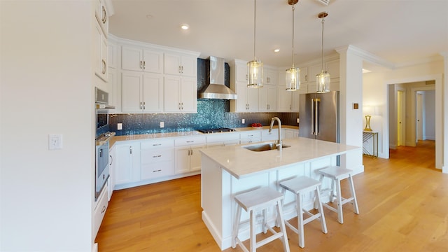 kitchen featuring sink, stainless steel appliances, wall chimney range hood, an island with sink, and white cabinets