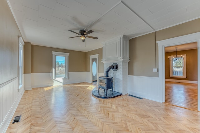 unfurnished living room featuring light parquet floors, a wood stove, ceiling fan with notable chandelier, and ornamental molding