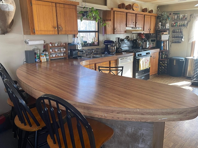 kitchen featuring white dishwasher, under cabinet range hood, a sink, brown cabinets, and gas range