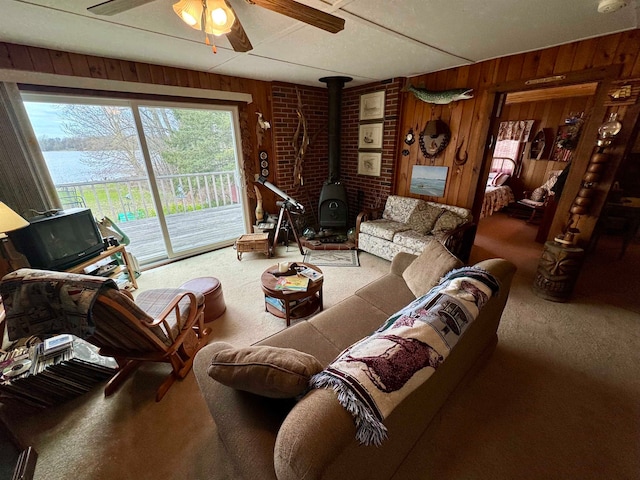 carpeted living room featuring a wood stove, ceiling fan, brick wall, and wooden walls