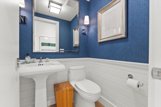 bathroom featuring a wainscoted wall, toilet, wallpapered walls, and a sink