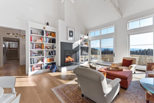 living room with a warm lit fireplace, ceiling fan, high vaulted ceiling, wood finished floors, and stairway