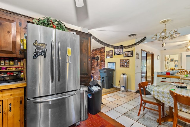 kitchen featuring light tile flooring, ceiling fan with notable chandelier, brick wall, hanging light fixtures, and stainless steel refrigerator