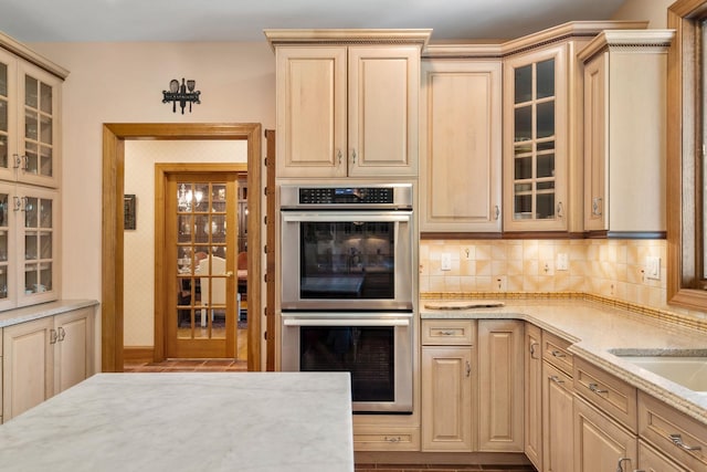 kitchen with light stone countertops, light brown cabinets, stainless steel double oven, glass insert cabinets, and backsplash