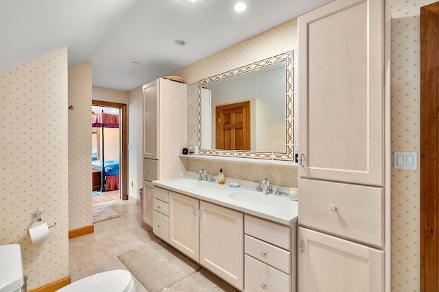 ensuite bathroom with wallpapered walls, double vanity, a sink, toilet, and connected bathroom