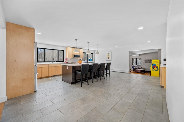 kitchen with a breakfast bar area, a kitchen island with sink, stainless steel appliances, light brown cabinetry, and decorative light fixtures