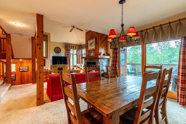 dining area with light carpet, ceiling fan with notable chandelier, a stone fireplace, and wood walls