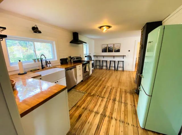 kitchen featuring stainless steel appliances, sink, butcher block counters, light wood-type flooring, and wall chimney exhaust hood