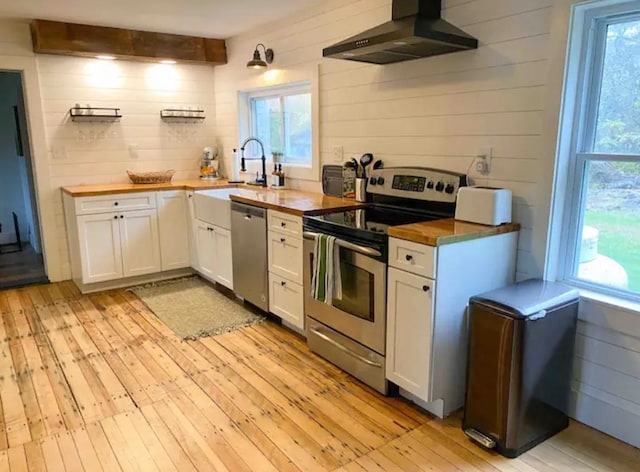 kitchen featuring butcher block countertops, appliances with stainless steel finishes, wall chimney range hood, light wood-type flooring, and white cabinetry