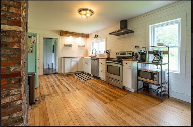 kitchen with white cabinetry, wall chimney range hood, light wood-type flooring, wood walls, and appliances with stainless steel finishes