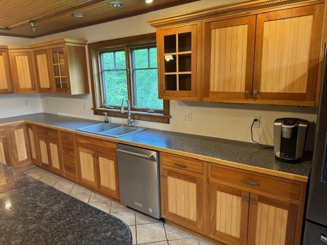 kitchen with dark countertops, glass insert cabinets, stainless steel dishwasher, a sink, and light tile patterned flooring