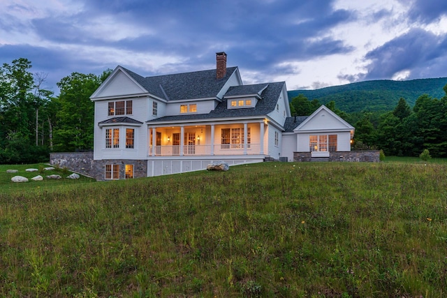 back of house with covered porch, stone siding, a chimney, a shingled roof, and a yard