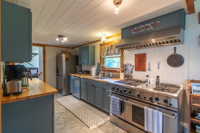 kitchen featuring appliances with stainless steel finishes, butcher block countertops, wood walls, sink, and wall chimney exhaust hood