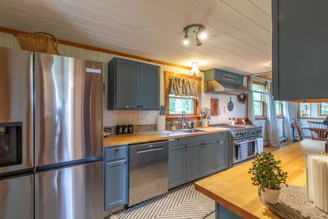 kitchen with appliances with stainless steel finishes, wood counters, sink, plenty of natural light, and wall chimney range hood