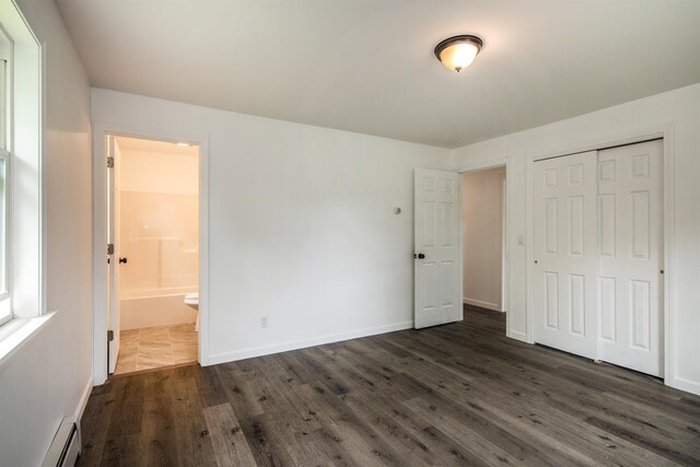 unfurnished bedroom featuring baseboards, dark wood finished floors, ensuite bath, baseboard heating, and a closet