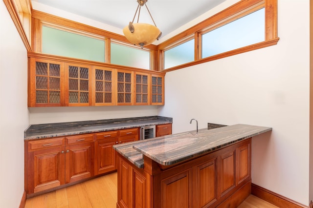 kitchen with dark stone countertops, kitchen peninsula, pendant lighting, beverage cooler, and light hardwood / wood-style floors