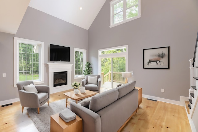 living room featuring high vaulted ceiling, light wood-type flooring, a glass covered fireplace, and visible vents