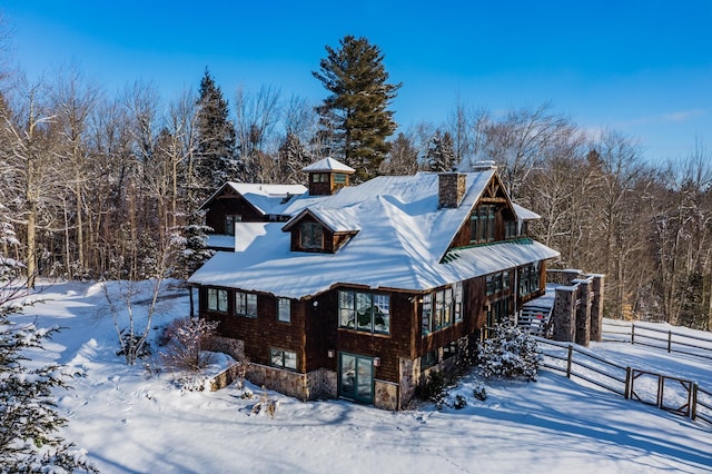 view of snowy exterior featuring stone siding, a chimney, a forest view, and fence