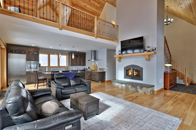 living area featuring high vaulted ceiling, a notable chandelier, stairway, light wood-type flooring, and a glass covered fireplace