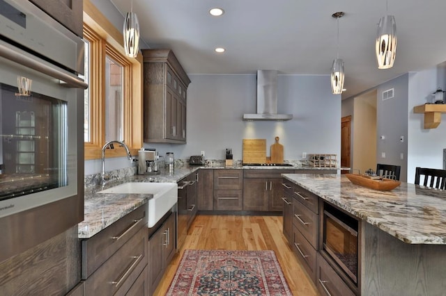 kitchen with stainless steel appliances, a sink, light wood-style floors, wall chimney exhaust hood, and decorative light fixtures