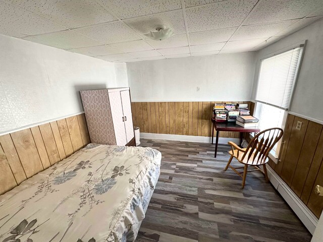 bedroom with a baseboard heating unit, wooden walls, dark hardwood / wood-style flooring, and a drop ceiling