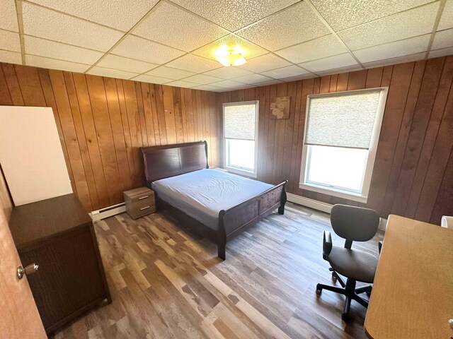 bedroom with baseboard heating, wood-type flooring, wooden walls, and a drop ceiling