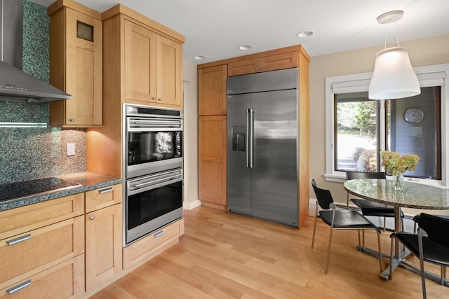 kitchen featuring hanging light fixtures, wall chimney range hood, backsplash, light hardwood / wood-style floors, and appliances with stainless steel finishes