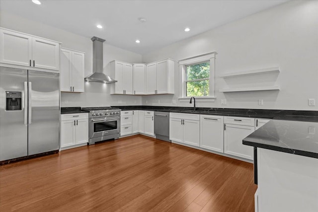 kitchen featuring wall chimney range hood, white cabinets, dark countertops, appliances with stainless steel finishes, and a sink