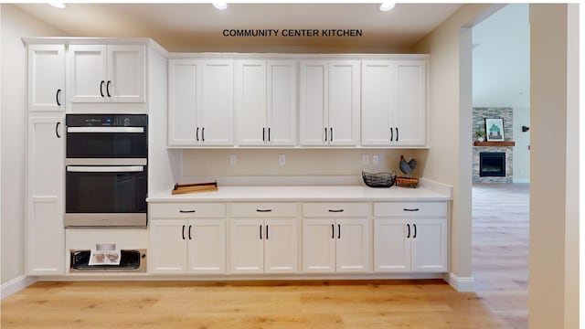 kitchen with a stone fireplace, white cabinets, stainless steel double oven, and light wood-type flooring