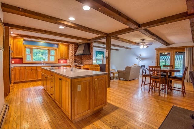 kitchen with light hardwood / wood-style floors, wall chimney exhaust hood, a kitchen island with sink, and ceiling fan