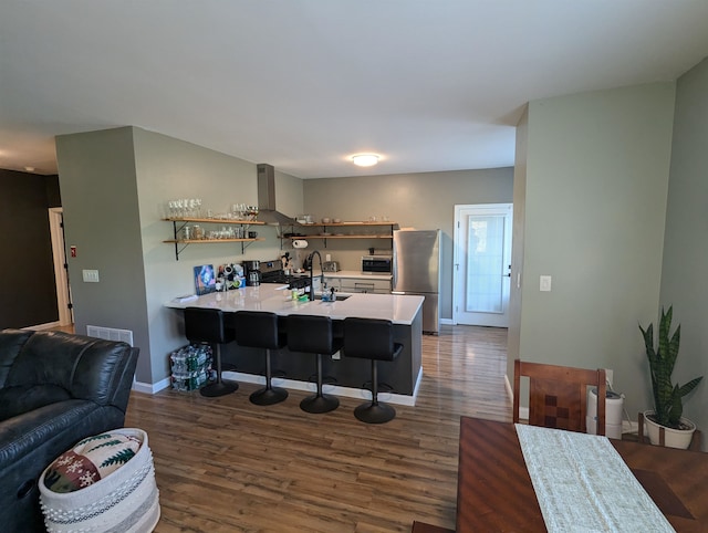 kitchen with dark wood-type flooring, kitchen peninsula, a kitchen bar, wall chimney exhaust hood, and stainless steel appliances