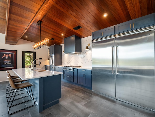 kitchen with wall chimney exhaust hood, sink, stainless steel appliances, hanging light fixtures, and an island with sink