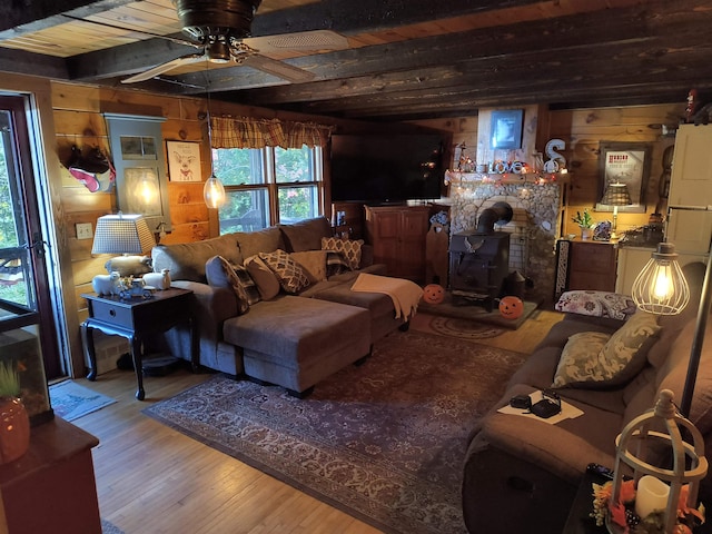 living room featuring ceiling fan, wood walls, beam ceiling, a wood stove, and hardwood / wood-style floors