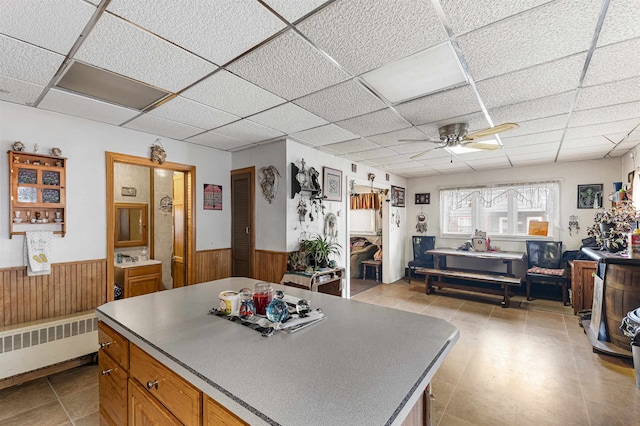 kitchen with ceiling fan, radiator heating unit, a center island, a drop ceiling, and wood walls