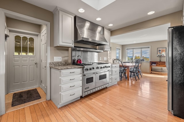 kitchen featuring white cabinetry, light hardwood / wood-style flooring, dark stone countertops, wall chimney exhaust hood, and stainless steel appliances