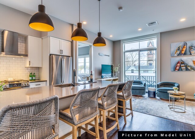 kitchen with stainless steel appliances, wall chimney exhaust hood, decorative light fixtures, wood-type flooring, and white cabinets