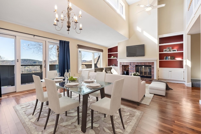 dining space featuring ceiling fan with notable chandelier, wood-type flooring, a fireplace, built in features, and a high ceiling