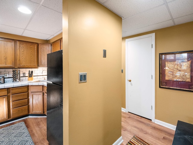 kitchen with a paneled ceiling, black fridge, backsplash, tile counters, and light wood-type flooring