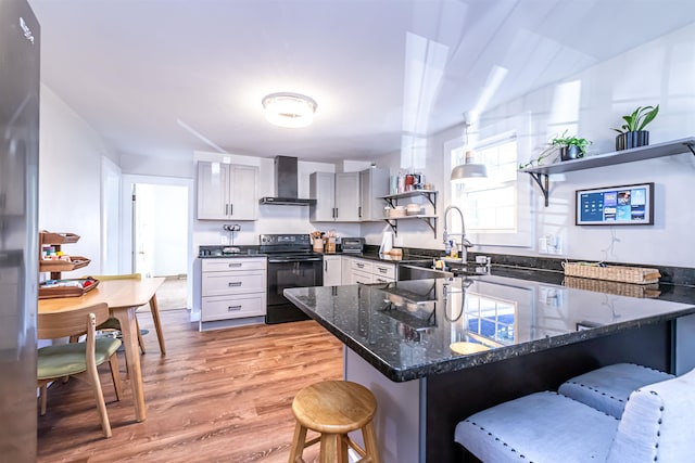 kitchen with wall chimney exhaust hood, a kitchen breakfast bar, light wood-type flooring, and black / electric stove