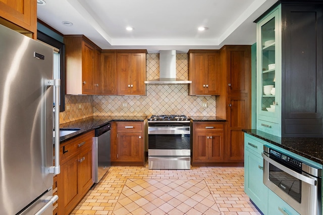 kitchen with wall chimney exhaust hood, a raised ceiling, appliances with stainless steel finishes, and dark stone counters