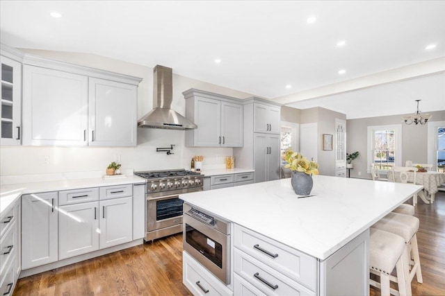 kitchen with appliances with stainless steel finishes, wall chimney exhaust hood, an inviting chandelier, hardwood / wood-style floors, and a breakfast bar area