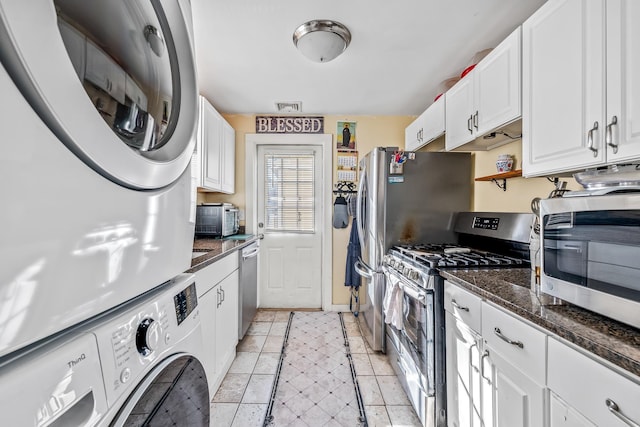 kitchen featuring white cabinets, stacked washer / dryer, appliances with stainless steel finishes, and dark stone counters