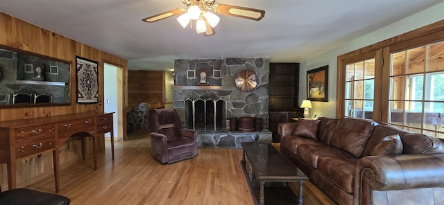 living room featuring light wood-type flooring, a stone fireplace, ceiling fan, and wood walls