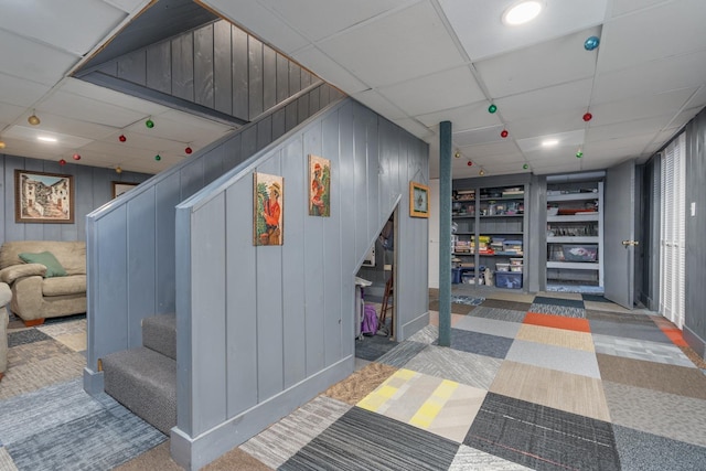 corridor featuring built in features, wood walls, a drop ceiling, and carpet