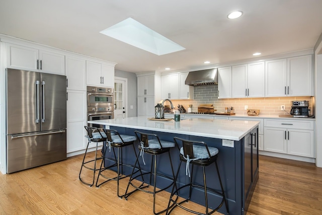 kitchen featuring white cabinetry, a skylight, a center island with sink, appliances with stainless steel finishes, and wall chimney range hood