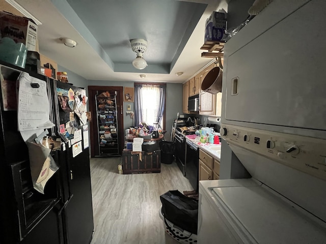 kitchen with stacked washer / drying machine, a tray ceiling, black fridge, light wood-type flooring, and dishwashing machine