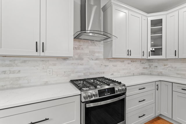 kitchen featuring light stone countertops, stainless steel gas stove, wall chimney range hood, decorative backsplash, and white cabinets