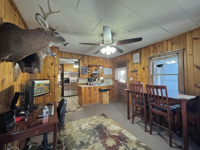 kitchen featuring ceiling fan, stainless steel fridge with ice dispenser, kitchen peninsula, light colored carpet, and wooden walls